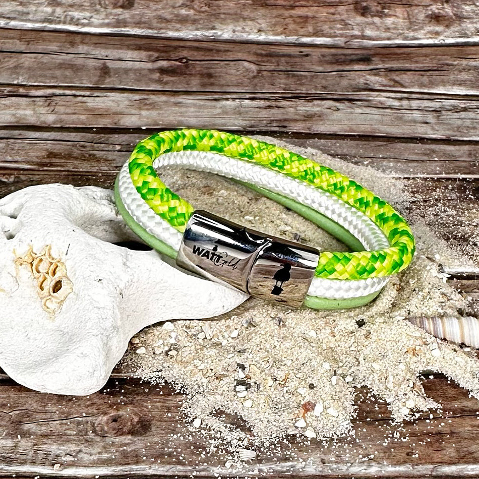 Ein grün-weiß geflochtenes Frühlingsbrise-Armband von WattGold mit silbernem Verschluss ruht auf einer mit Muscheln und Sand verzierten Holzoberfläche und fängt die Essenz seines Namens wunderschön ein.