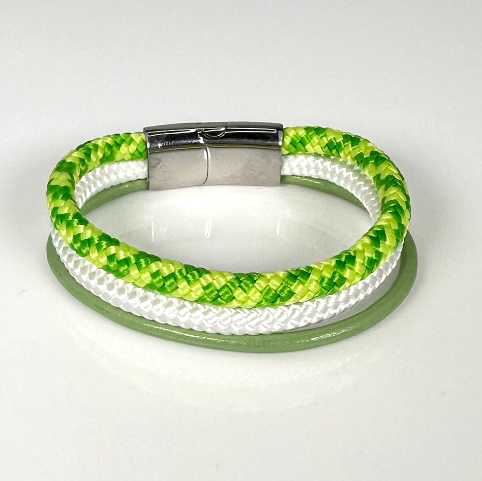 Die Frühlingsbrise von WattGold ist ein gewebtes Armband aus leuchtend grünen, weißen und gelben Strängen, kombiniert mit einem glänzenden silbernen Verschluss. Es ist elegant in einer kreisförmigen Form auf einer reflektierenden weißen Oberfläche angeordnet.